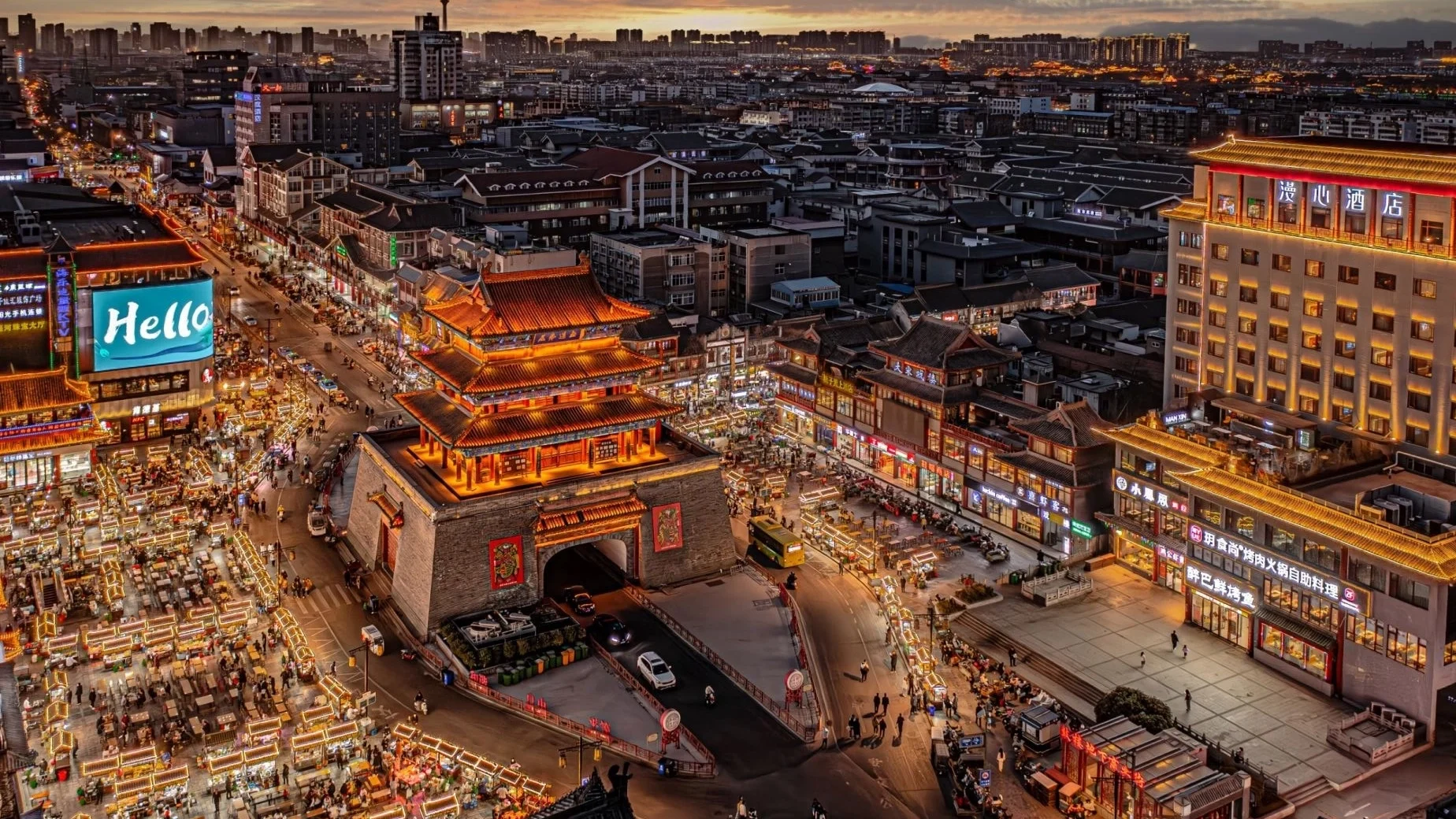 Luchtfoto van de bruisende Drum Tower van Kaifeng en de omliggende marktstraten, verlicht in de schemering, die een levendig stadsleven en historische architectuur laten zien