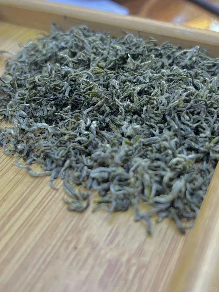 A close up of dried Duyun Maojian tea leaves with a slightly curled texture spread on a wooden tray