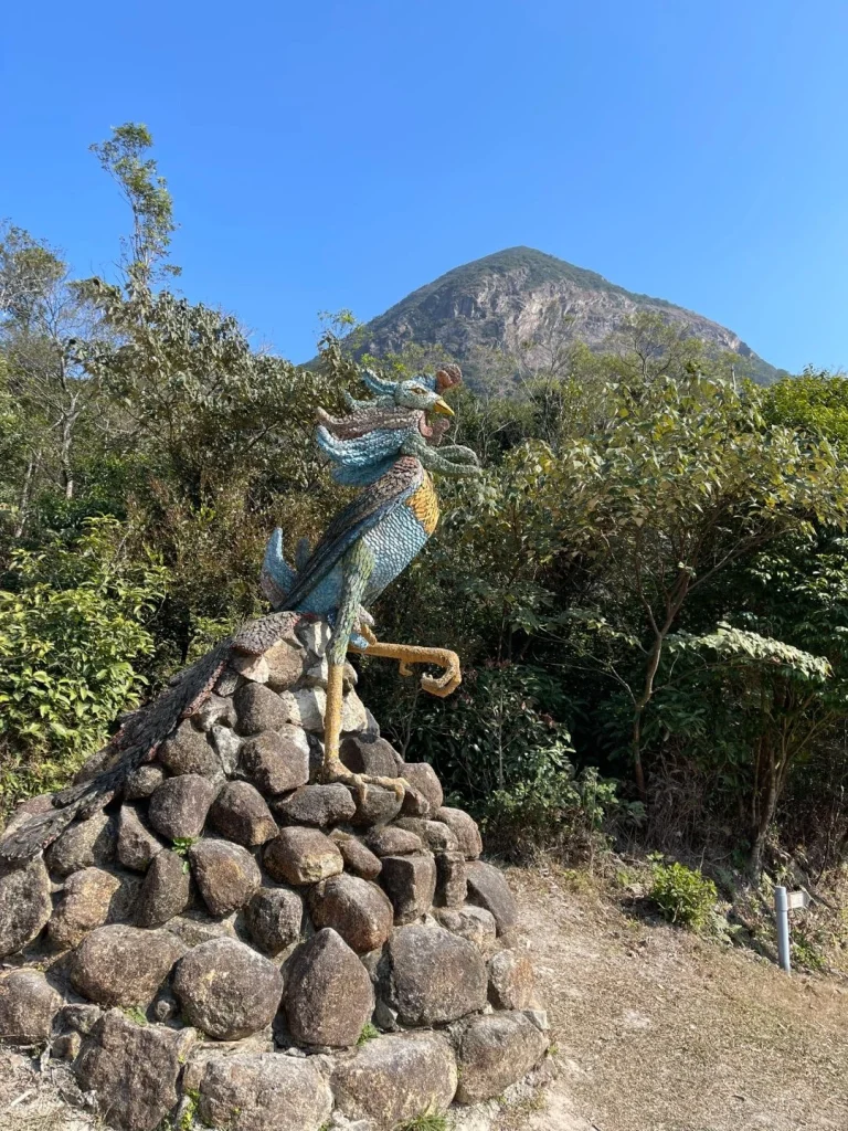 A colorful mosaic sculpture of a rooster in the natural landscape of Hong Kong with mountains in the background