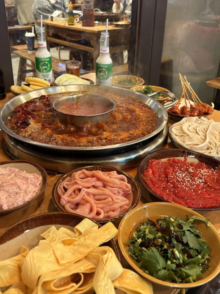 A vibrant Sichuan hotpot surrounded by fresh meats vegetables and sauces with an aromatic spicy broth