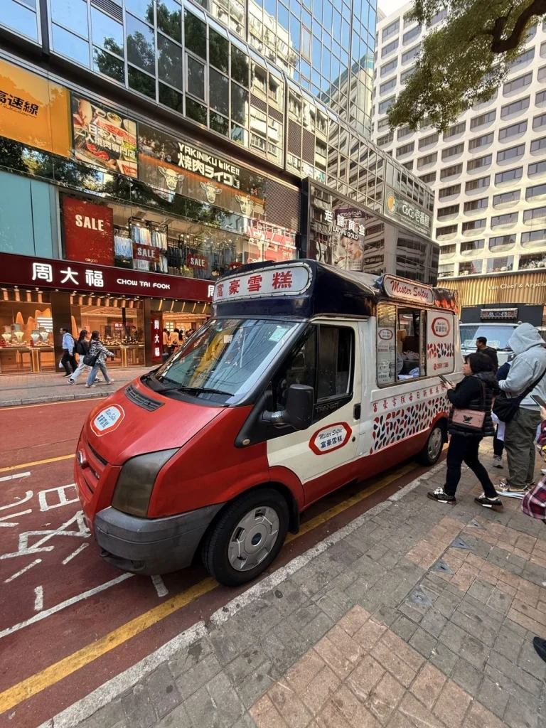 An ice cream truck parked in the busy streets of Hong Kong with shops and bright signage in the background
