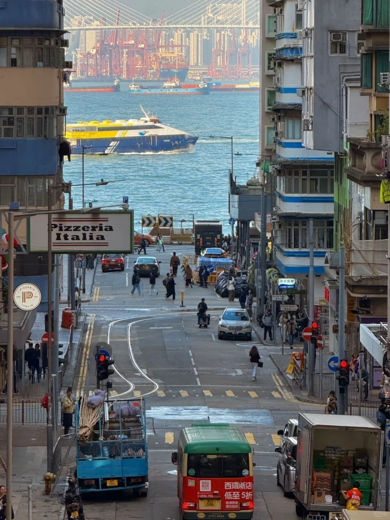 Busy Hong Kong street with vehicles and sea view View of a bustling Hong Kong street with busy traffic shops and a ferry passing in the background