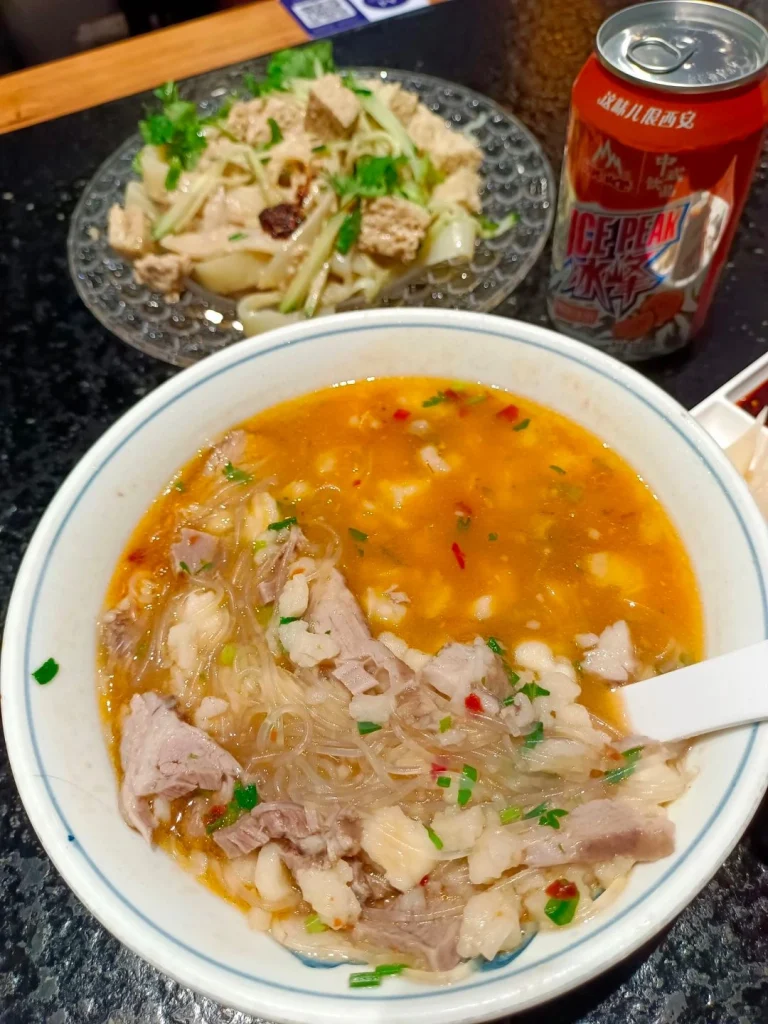 Xian Restaurant dishes A bowl of Xian noodle soup with meat accompanied by a plate of tofu salad and a can of Ice Peak soda