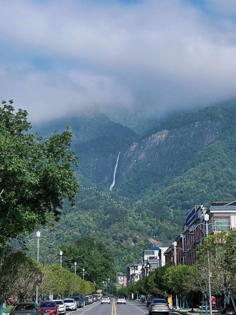 Lushan Waterfall in the Mist Tall waterfall flows from cloud covered mountains into the valley above a vibrant town in Lushan
