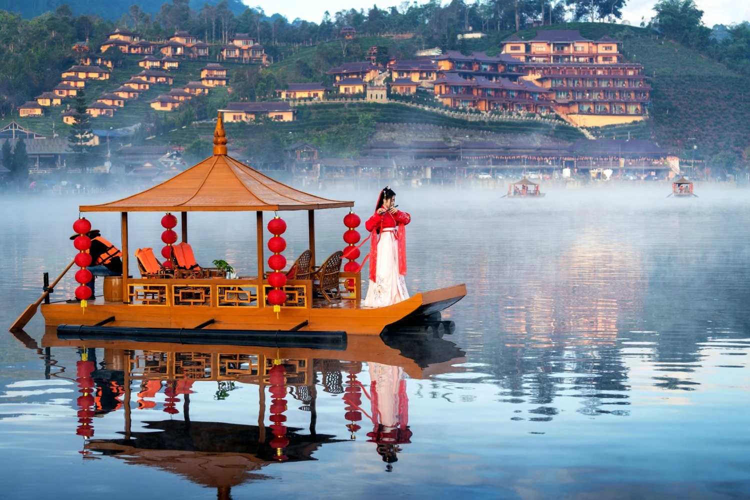 Barca tradizionale cinese con lanterne rosse su un lago nebbioso, donna in hanfu che suona il flauto, ville sulle colline sullo sfondo: scenario di un viaggio culturale in Cina.