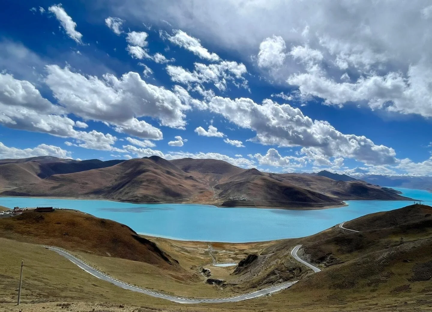 Vista aérea del lago Yamdrok con una sinuosa carretera de montaña sobre las aguas turquesas, colinas onduladas y nubes espectaculares - Guía de viaje al Tíbet: mejores rutas, consejos e itinerarios 2026 - The China Journey Vista aérea del lago Yamdrok con una sinuosa carretera de montaña sobre las aguas turquesas, colinas onduladas y nubes espectaculares - Guía de viaje al Tíbet: mejores rutas, consejos e itinerarios 2026 - The China Journey