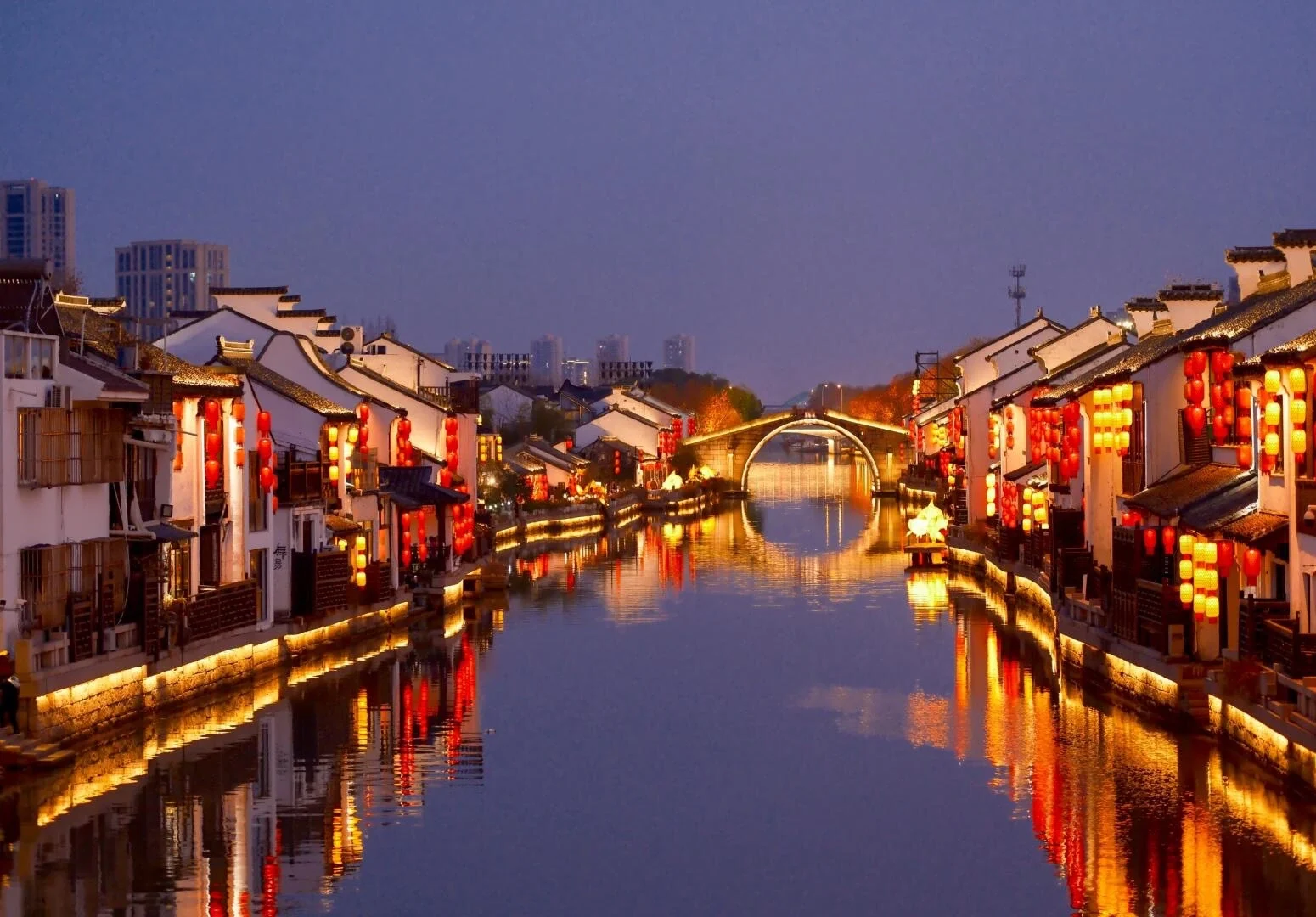 Kanal med lanterner om aftenen i en Jiangnan-by i Wuxi, traditionelle huse, røde lanterner og hvælvet bro spejlet i vandet med festlig stemning - Wuxi rejseguide: Bedste rejseplan og tips 2026 - The China Journey Kanal med lanterner om aftenen i en Jiangnan-by i Wuxi, traditionelle huse, røde lanterner og hvælvet bro spejlet i vandet med festlig stemning - Wuxi rejseguide: Bedste rejseplan og tips 2026 - The China Journey